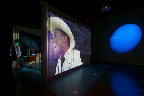Projection on a large curtain of a man wearing a white cowboy hat mid-speech. A set of a play is behind the curtain and a large blue spotlight on the right wall lights the gallery.&nbsp;