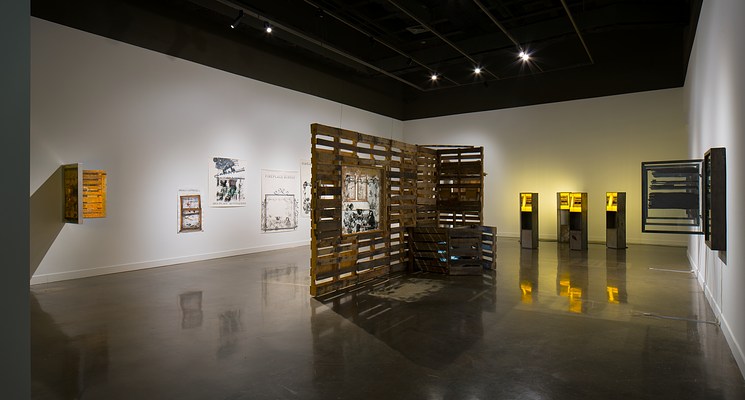 Installation view with a large sculpture in the center of the room made of wood pallets. There are prints on the wall to the left and sculptures that incorporate light on the right.&nbsp;
