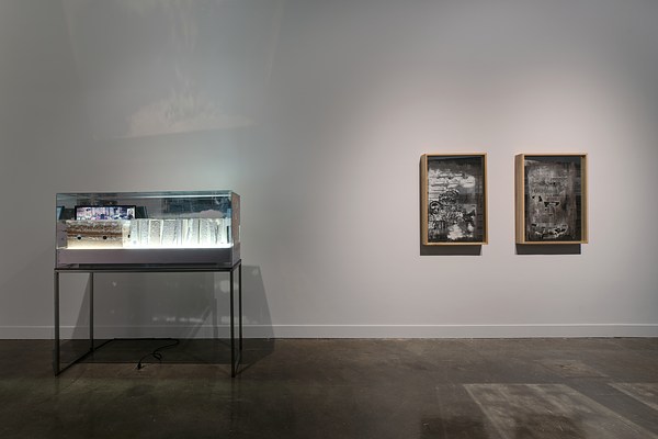 Large glass box on a podium with light and paper encased inside and two framed gray prints on the right wall.&nbsp;