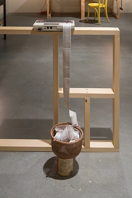 A small computer/ printer on a ledge, paper spooled over into a woven basket on top of a paint can, a lighted lamp in back.