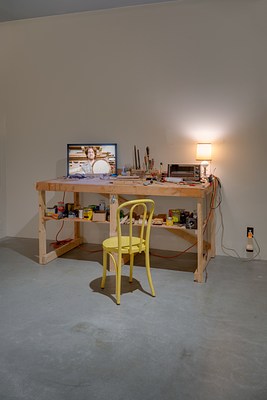 A work bench with a monitor, tools, a toaster oven, a lamp with a warm glow, and a yellow wood chair in front.