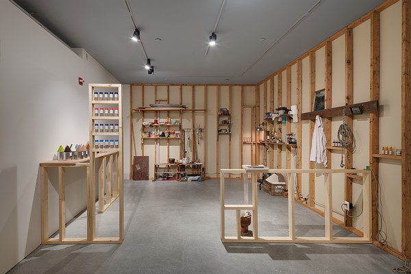 Installation view of a space with wood framework, shelves with objects, a jacket, rope, computer, a spice rack with bottles.