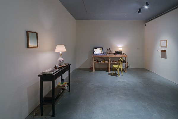 A space with workbench, lamp and chair in back, a picture and shelf on the right, a mirror and table with a lamp on the left.