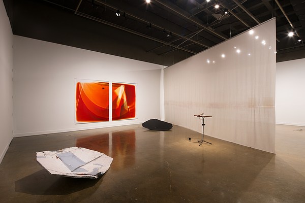 A large space bisected with hanging fabric, two large orange prints on a wall, a wooden sound piece, and two floor sculptures.
