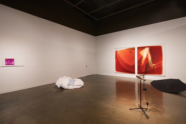 A large space bisected with hanging fabric, two large orange prints on a wall, a wooden sound piece, and two floor sculptures.