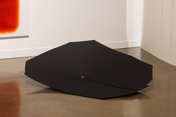 On the floor between a fabric hanging piece and a color print is a large black shield piece made of posters, plastic, metal.
