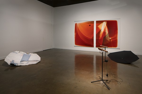 Two orange and white prints on a wall, a black and a white shield sculpture on the floor, a wood sound piece on a stand.