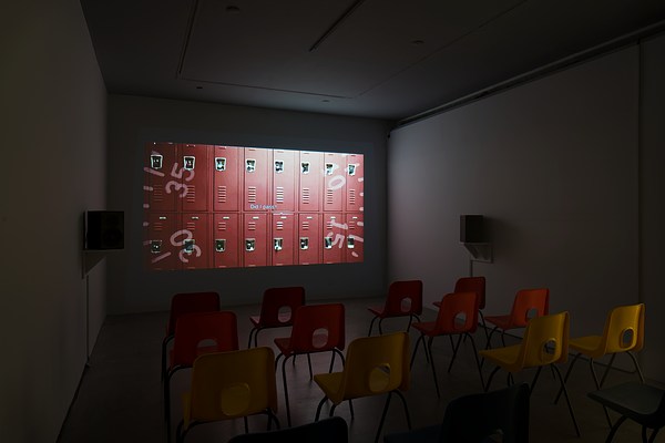 A darkened gallery space set up like a theater with speakers, chairs and a projected video showing two rows of red lockers