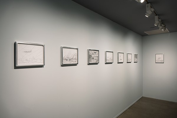 A wall on the left with seven framed drawings hung diagonally, and at the far end a perpendicular wall with one drawing