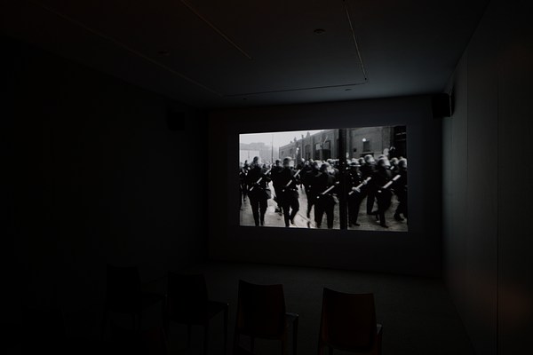 A black and white video of a group of police wearing helmets with face shields and clutching batons, displayed in a dark room.