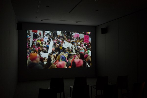 A colorful video scene of a large crowd of protesters, many holding signs, plays on a screen in a dark room.