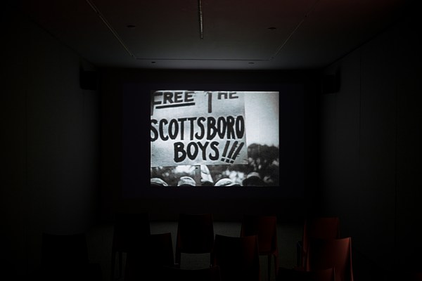 In the center of a dark room, a black and white video displays a protest sign that says “Free the Scottsboro Boys”.