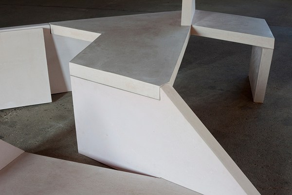 Close up view of a concrete bench sculpture, made of geometric shapes with a triangle sticking up from the seat area, sits on a gray floor.. 