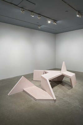 A performance piece in a gallery where a person wearing navy squats lies face down on a bench sculpture, arms stretched falling over the top.&nbsp;