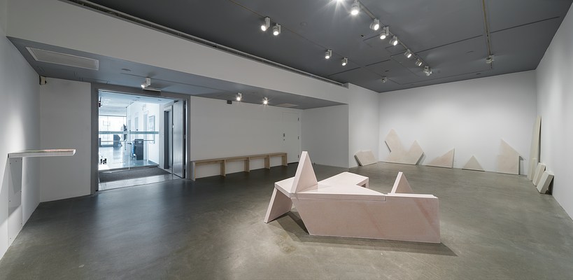 A concrete geometric bench sculpture sits in the center of the gallery and irregularly shaped cast concrete panels of differing sizes line the walls in the background.
