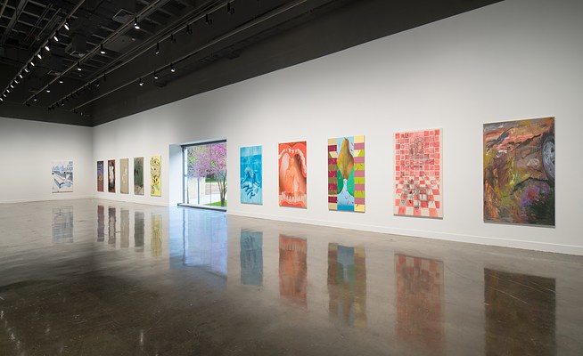 Ten colorful paintings hang side by side, a large window in the middle. A painting hangs on the far intersecting wall.