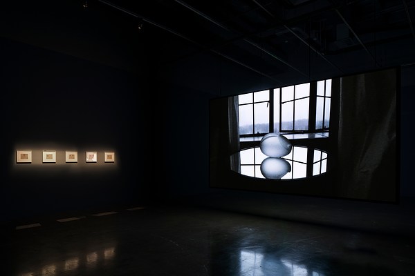 Wide shot of dark room with 5 illuminated artworks on the left, a silvery gray image projected on a screen to the right.