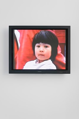Video monitor shows a closeup of a young girl with short black hair staring at the viewer, background saturated in red.