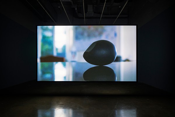 Video of black ovoid &amp; its reflection, with blurry blue cast bac k ground projected on s creen in dark room.
