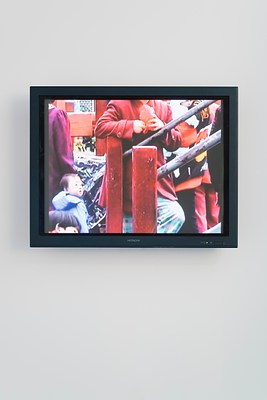 Video monitor shows a colorful scene of a torso in a red coat behind a railing, a small child, glimpses of others around them.