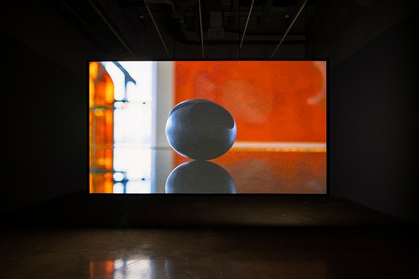 Video of black oval object with reflection, in a diffusely lit orange space, is projected on a hanging scre en in a dark room.