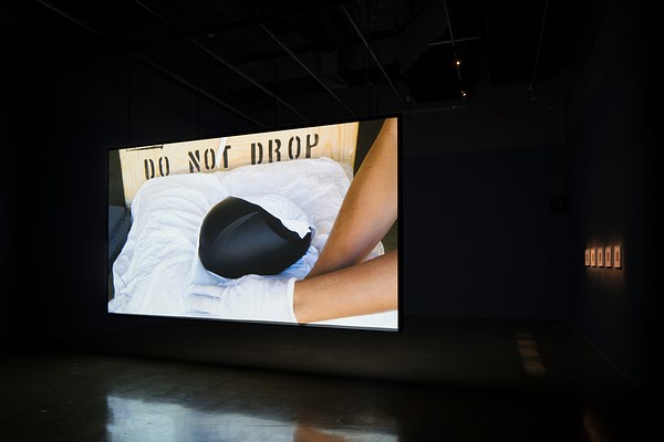 Video projection of a black object on a white pillow &amp; 2 arms with gloved hands. Do Not Drop is s tenciled on crate in back.