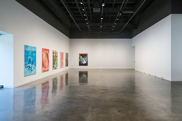 Long view of a gallery, six colorful paintings hang on the left, the right side is left blank except for words on a baseboard.