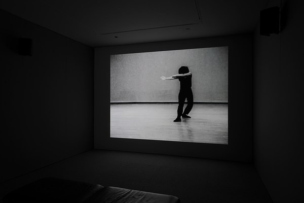 Video display of a dancer in black, posed with head bowed, both arms stretched out to her right, with right leg forward.