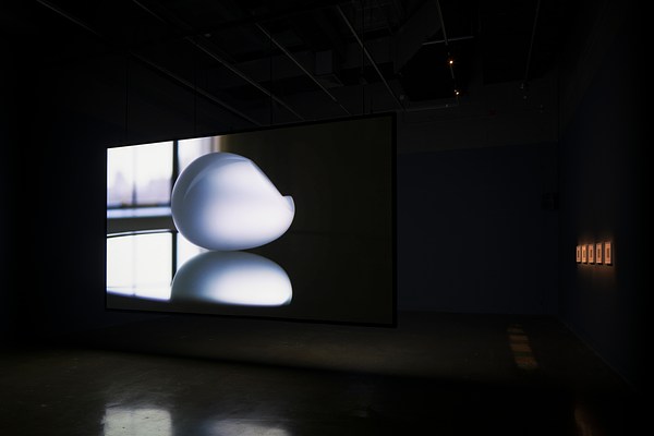 A silver gray oval shaped sculpture with reflection is project ed on a screen, 5 illuminated artworks on the right.