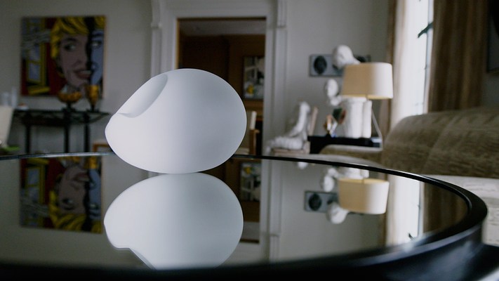 Video still of frosted crystal object on a mirrored table, reflecting its image and surrounding art filled room.