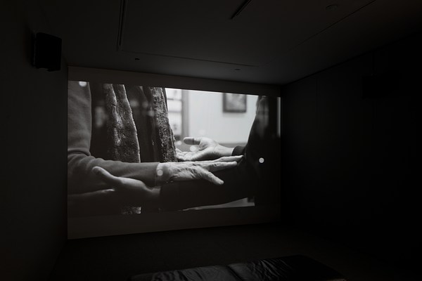  Dark room with video projection of a person's torso, arms bent at right angle, palms up, supporting the arms of another.