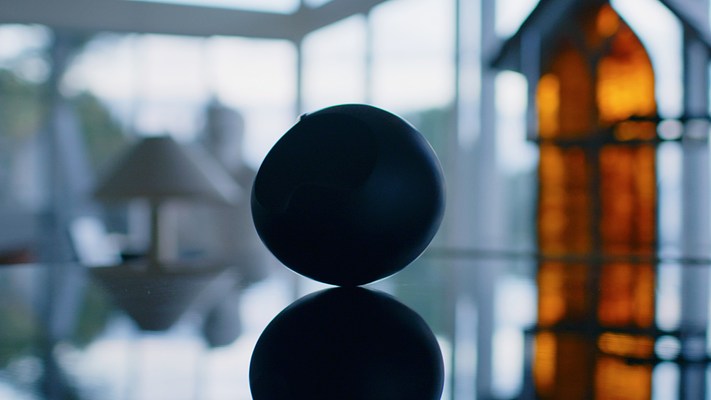 Video still of a black object on a mirror reflecting its image . Background is in soft foc us, and blends with reflection.