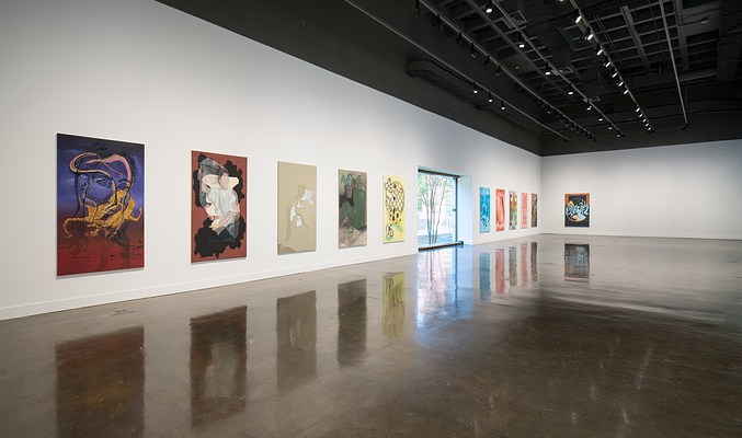 A long shot of a gallery with a series of paintings done in different palettes, a large window between them.