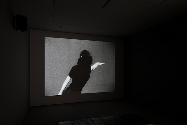Black and white video projected in a dark room displays woman in black from behind, stretching her right arm in a dance pose.