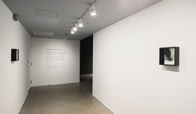 Corridor with track lights, small works on left and right walls. Wall of text at the far end leads into a darkened room.