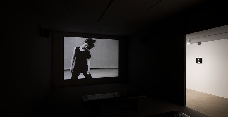 A dancer in motion is projected in a dark room. An opening leads to a bright hall where one small work hangs.