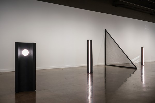 Three black standing sculptures, each with a different light, and a large black mesh  triangle jutting from the wall.