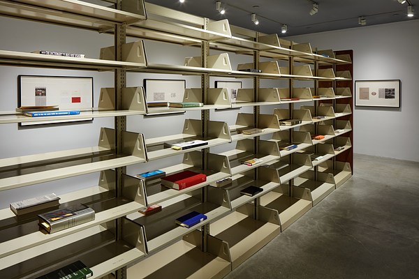 Long bookshelf stretches into room. Some shelves are empty, the rest each have one book face up, with 4 framed  works on walls.