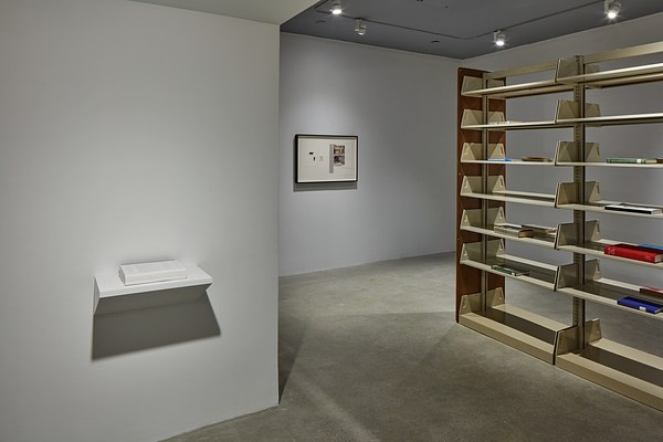 Three artworks including white shelf with white book to the left, a framed work hung in the center and a bookshelf to the right.