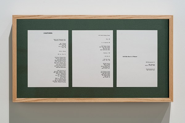  Hanging from a white wall, a wooden rectangle frame surrounds green matting and 3 pieces of paper  on which addresses are shown.