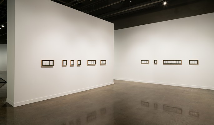  Nine horizontal frames with dark matting and white pieces of paper hang from two  nearly bisecting white walls.