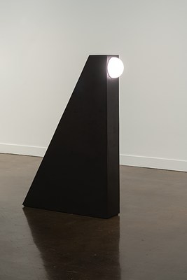 A black triangular tower stands alone in the Gallery. A large circular light protrudes and shines from the top right side.