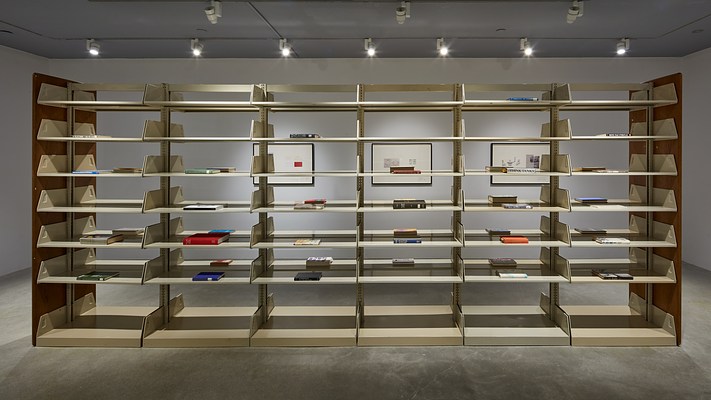 Metal library shelving fills the image. Single books with spines facing out lay on some shelves. Three framed works are visible beyond.&nbsp;