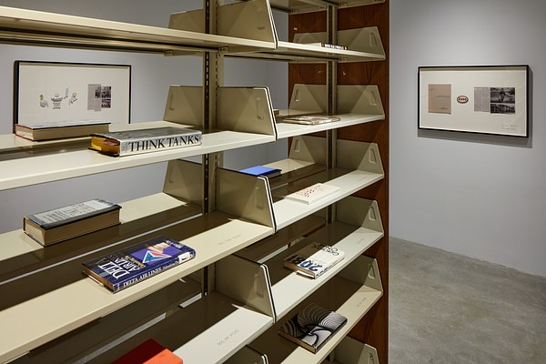 Metal bookshelves jutting from left, one book lying flat on each shelf, with 2 framed works on walls in background.