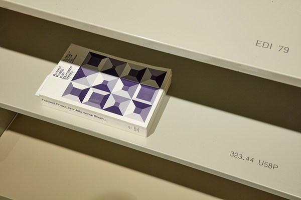 Closeup of 3 library shelves, book with purple pattern is face up on center shelf. Call numbers are  visible on top 2 shelves.