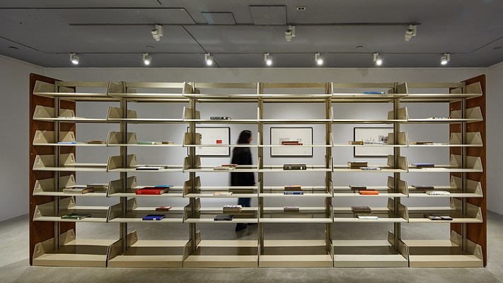 Metal library shelving fills the image. Single books with spines facing out lay on some shelves. Three framed works are visible beyond.