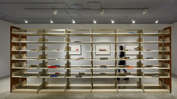 Metal library shelving fills the image. Single books with spines facing out lay on some shelves. 3 framed works  are visible beyond.