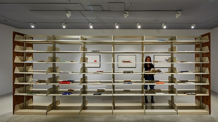 Metal library shelving fills the image. Single books with spines facing out lay on some shelves. 3 framed works  are visible beyond.