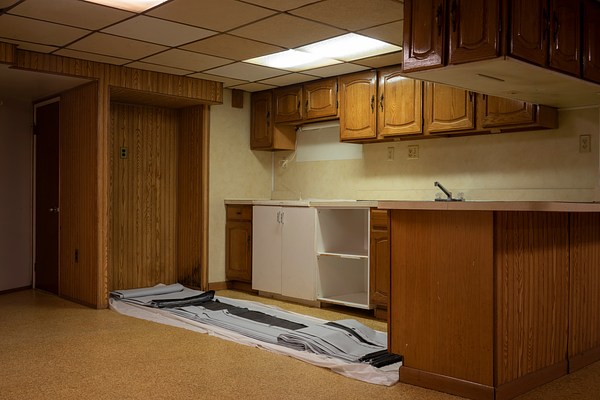 An installation view depicting a wood-paneled kitchen with yellow-brown wood cabinetry and no appliances. A printed banner is folded up into a long line, as if it was unrolled on the floor parallel to the cabinets. The scene is lit by a single fluorescent office light inset into a yellowed drop ceiling. The unrolled folded banner lays between a wood-paneled peninsula and an alcove for a missing refrigerator paneled with the same wood. The overall impression is surprisingly contrary to the expected look of a place where contemporary art is exhibited: dimly lit, covered in brown and tan toned surfaces, the fact that it's set up as a kitchen. The artwork is similarly surprising relative to traditional artworks: a piece of printed matter installed folded up on the floor.