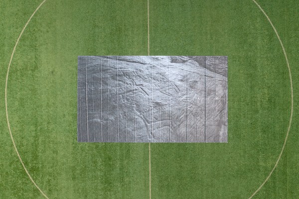 An ambiguous image that appears to depict a printed banner laid out flat on a green sports field emblazoned with white lines. Shot from above, both the field and banner are parallel to the picture plane and the banner, the same proportion of rectangle as the photograph itself, is positioned in the center. The so-called "banner" is recognizable as such only upon zooming in and seeing the grommets running along its edges. Printed on the banner is a grayscale image that is hard to read: the surface of the banner itself and the surface of whatever is printed on it: some kind of rough surface, maybe a sidewalk, with a corner of grass visible in its top right. Some kind of letters are scrawled in the surface but are illegible.